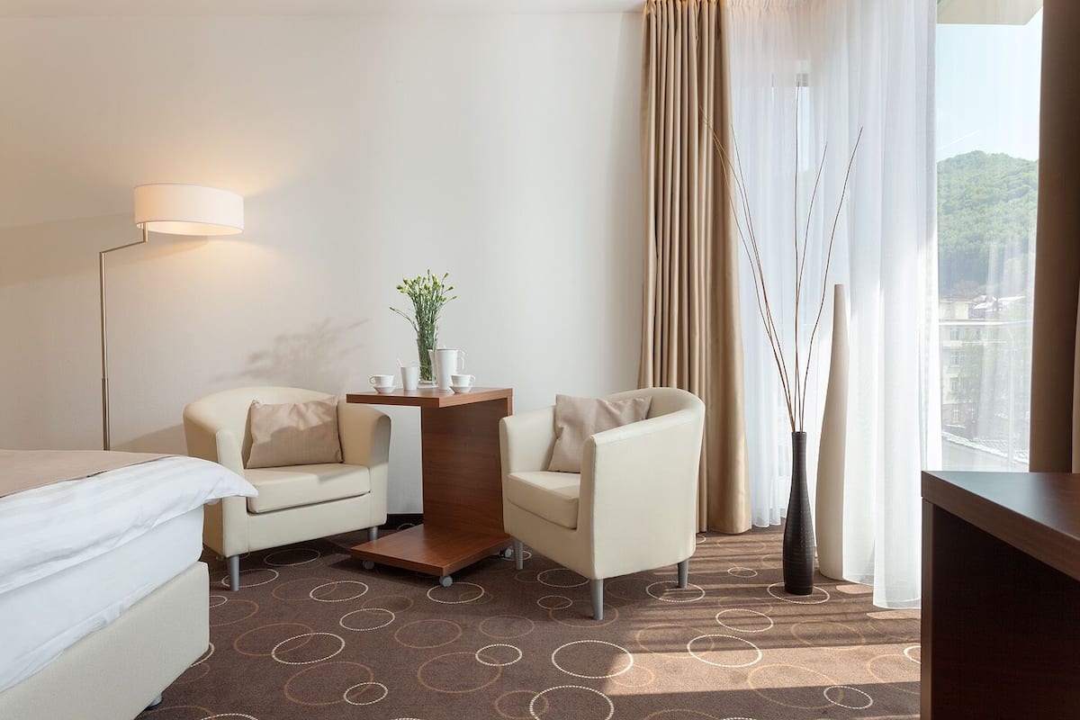 Quarto de hotel com uma cama, duas poltronas, uma pequena mesa de madeira com um vaso, um abajur e vista para a paisagem urbana através de cortinas finas.