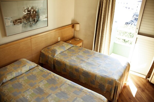 A hotel room with two beds, a bedside table with a lamp, and a window with curtains.