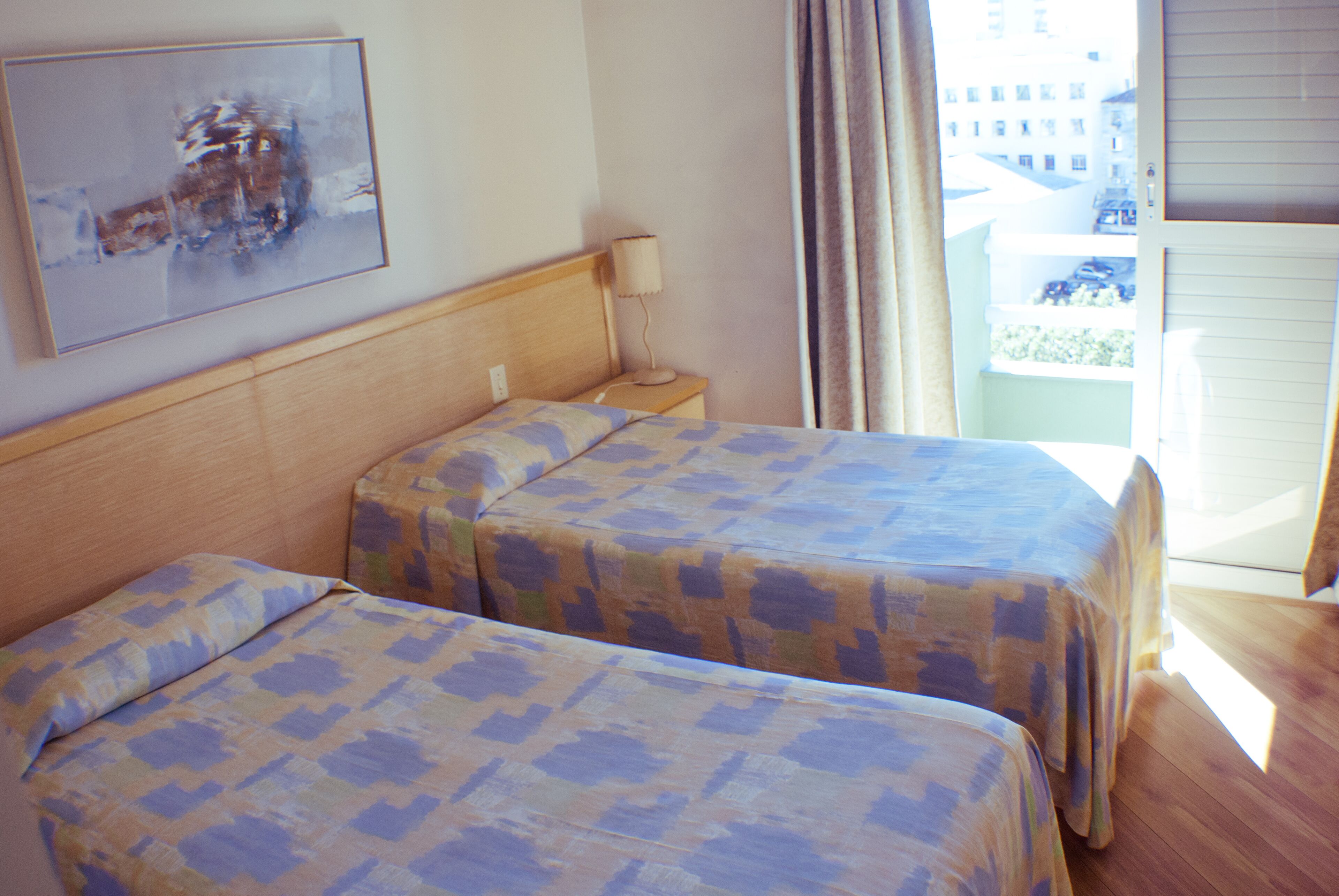 Two beds with patterned bedspreads, a bedside lamp, a painting on the wall, and a view of buildings through a window.