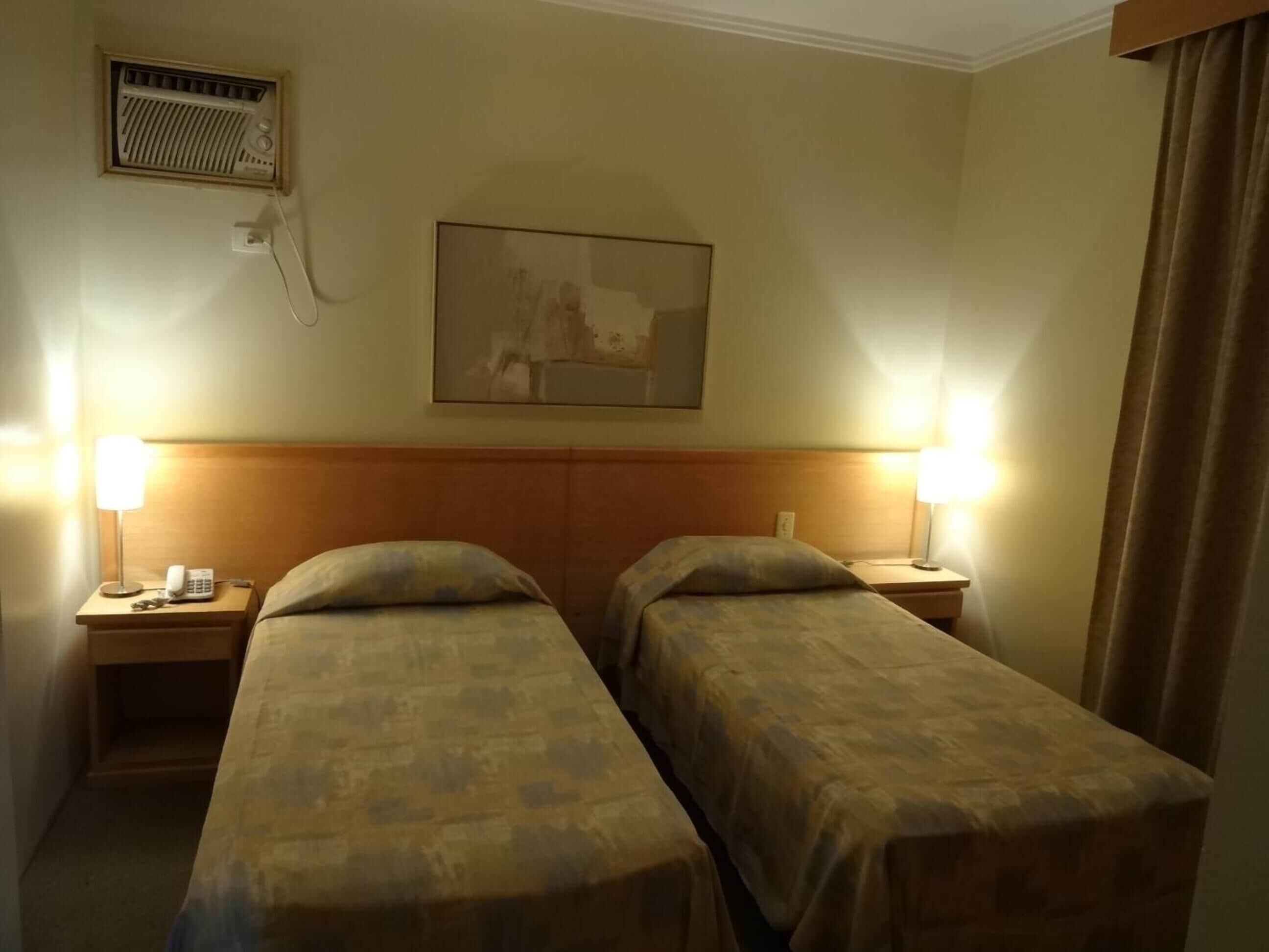 Een hotelkamer met twee eenpersoonsbedden, een nachtkastje met een lamp, een telefoon en een wandradiator.