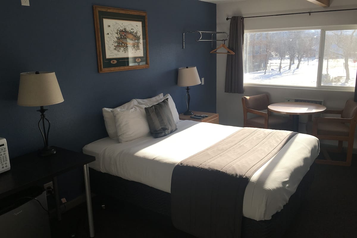 Une chambre d’hôtel équipée d’un lit, de lampes de chevet, d’un bureau, d’une chaise, d’une petite table et d’une fenêtre donnant sur un espace extérieur enneigé.