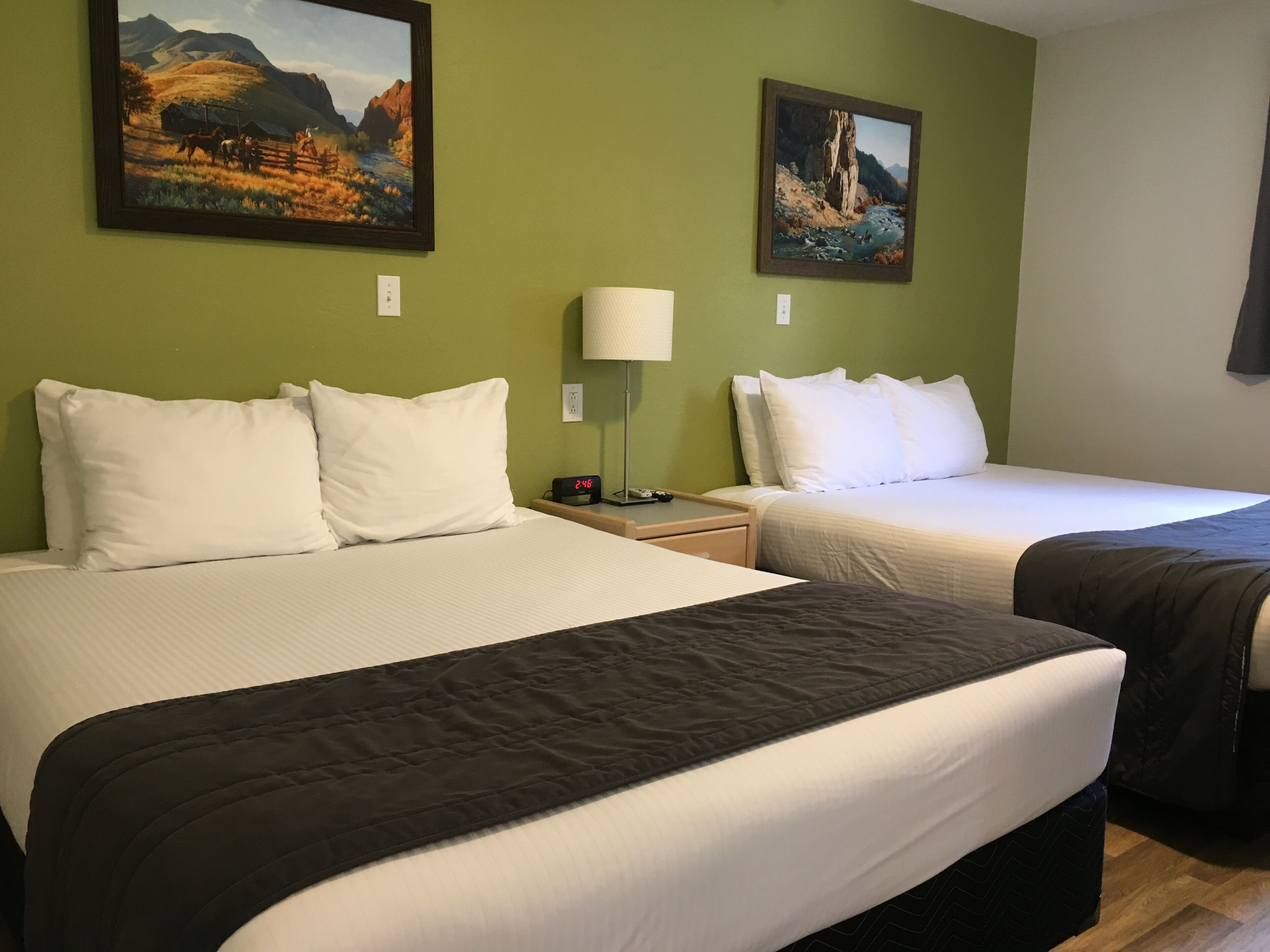A hotel room with two beds, a nightstand with a lamp, and two paintings on the wall.