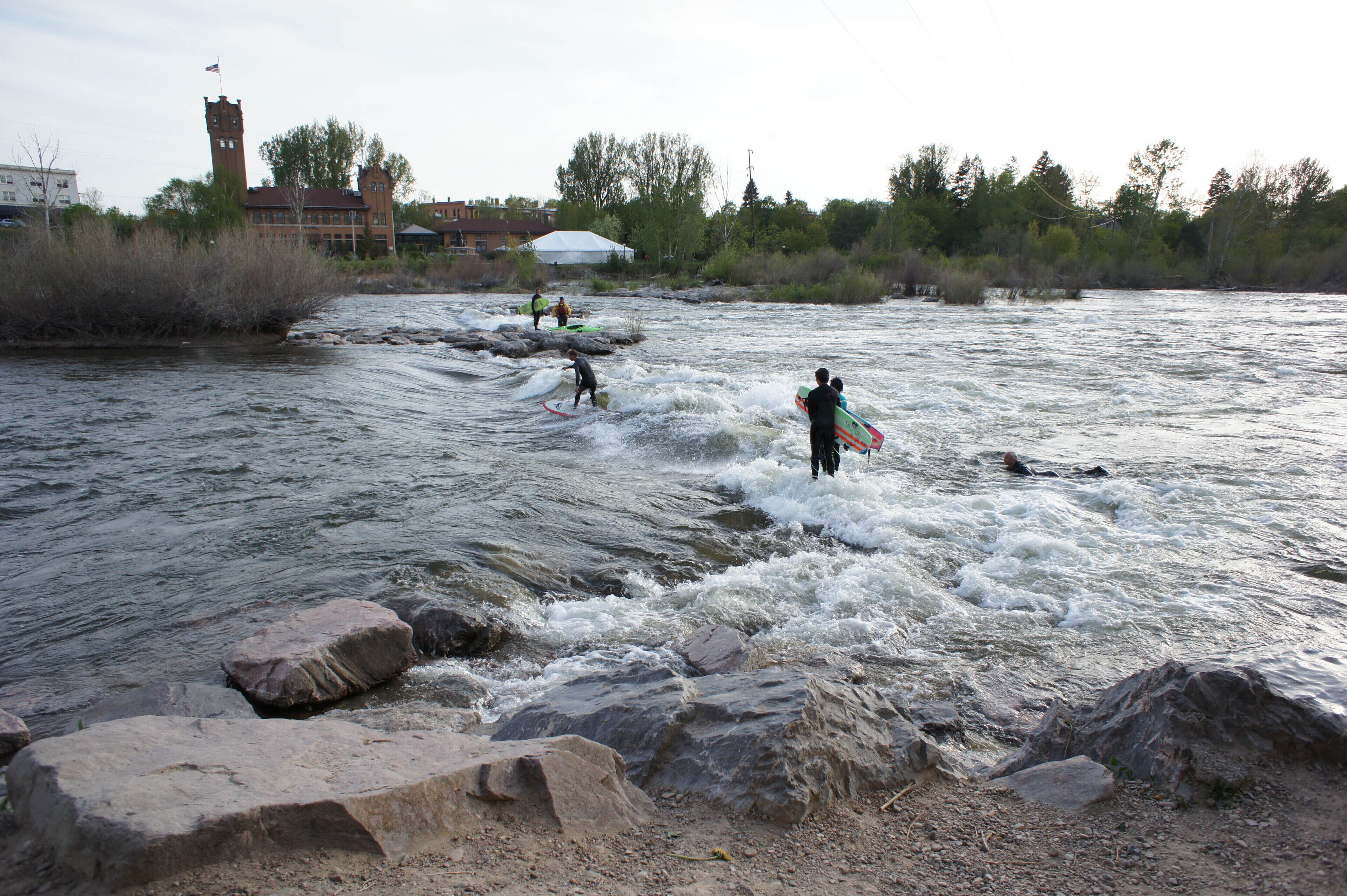 Un río con gente surfeando y remando. Hay rocas en el agua y un edificio con una torre al fondo.