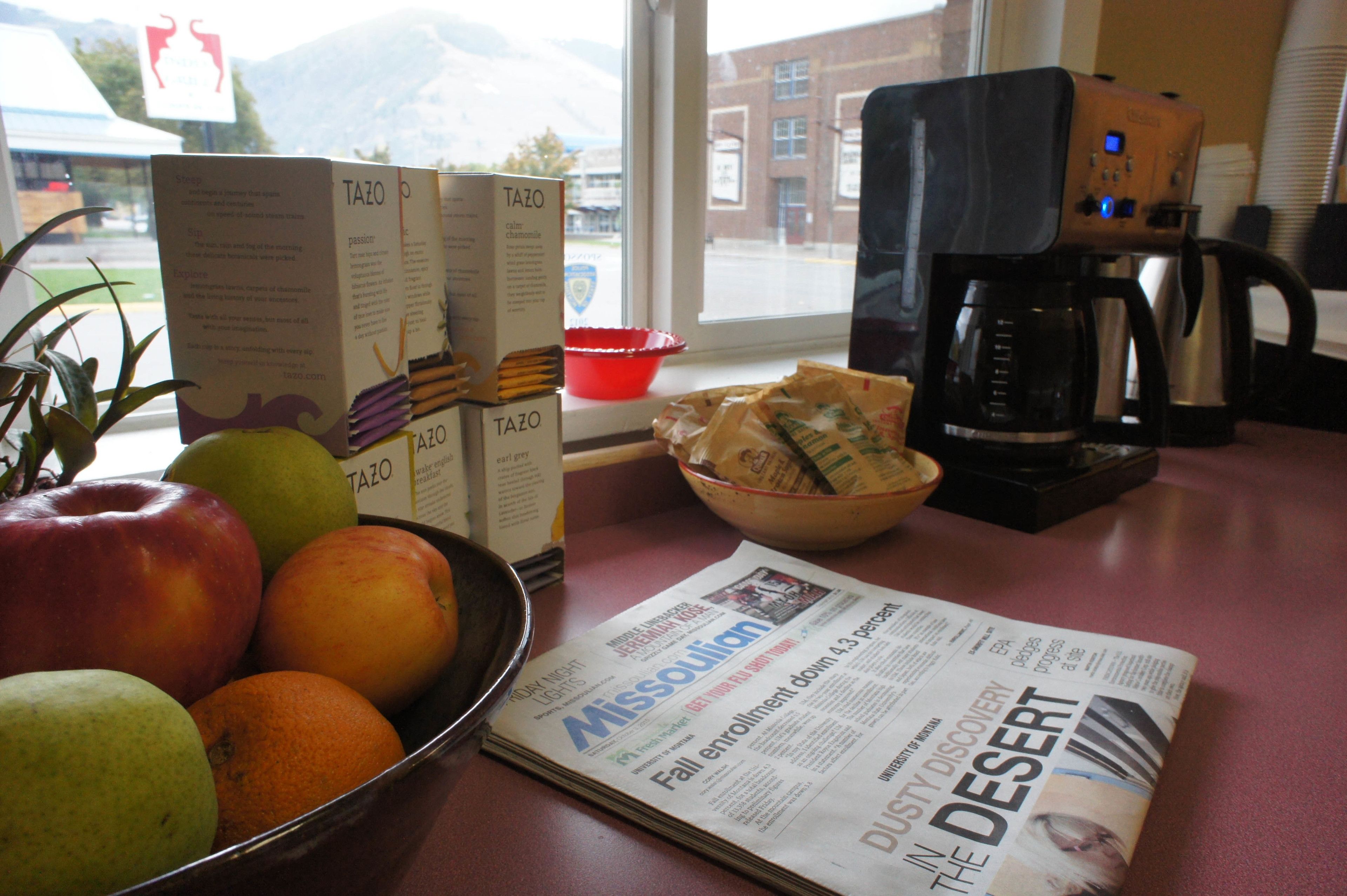 A table with a bowl of fruit, a coffee maker, a newspaper, and Tazo tea boxes.