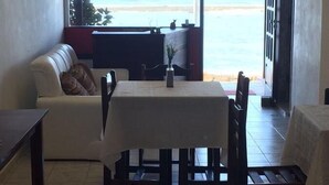Un área de comedor con una mesa y sillas, un sofá, y vista a la playa a través de amplias ventanas.