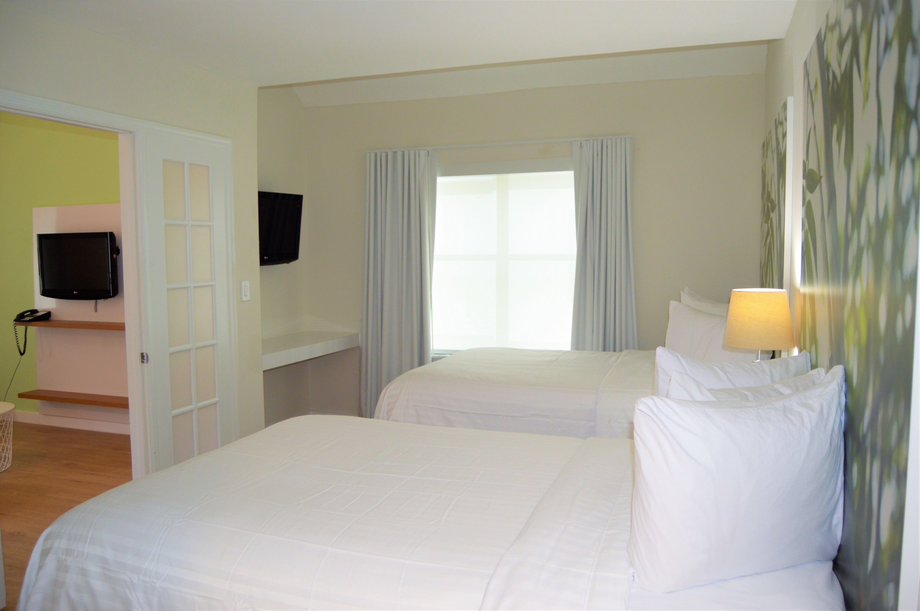 A hotel room with two beds, a TV mounted on the wall, a window with curtains, and a lamp on the bedside table.