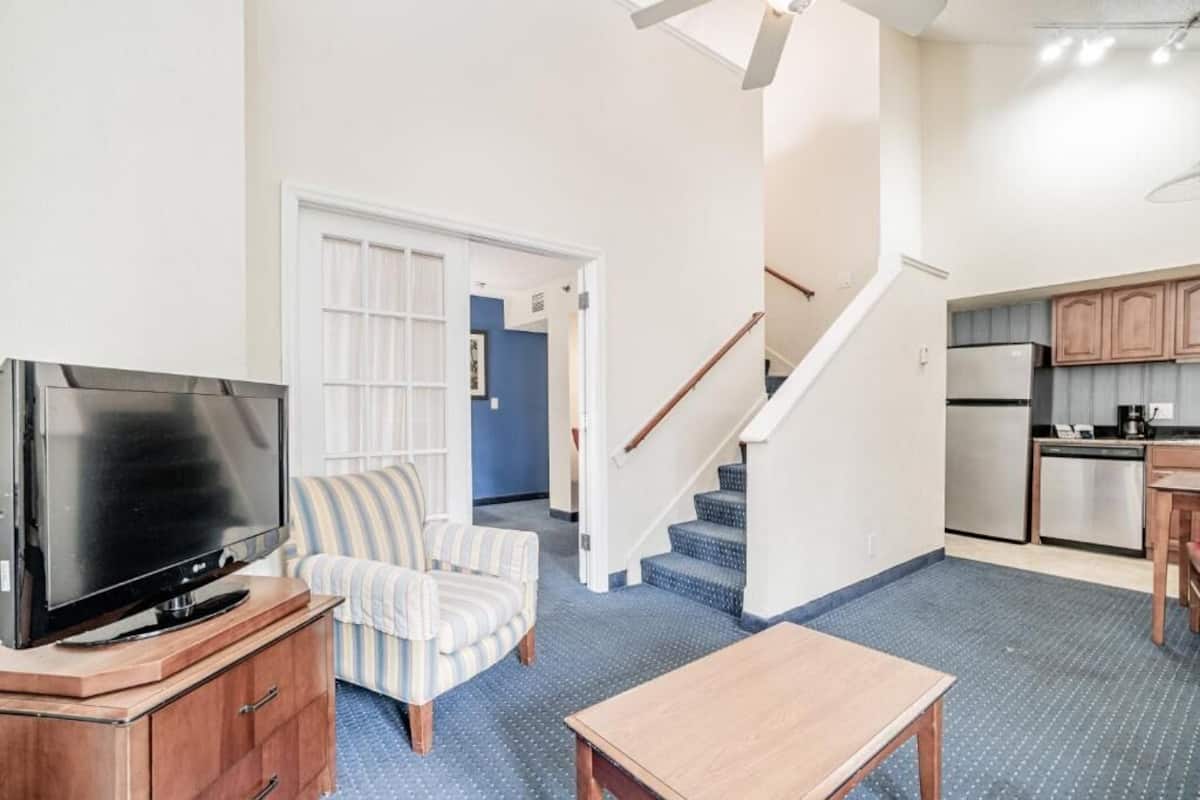 Een woonkamer met een televisie, een gestreepte fauteuil, een houten salontafel en een trap die naar een keuken leidt.