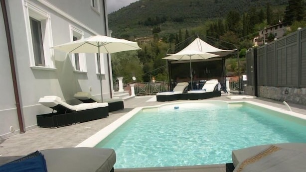 Piscina rectangular con sillas de descanso y sombrillas, rodeada por un muro de piedra y con una montaña al fondo.