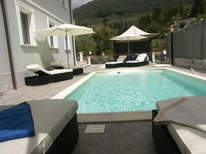 Ein rechteckiger Pool mit Liegestühlen und Sonnenschirmen, umgeben von einer Steinmauer und einem Bergpanorama.