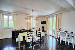 Un comedor con una larga mesa, sillas, una araña, un televisor y un piano.