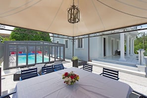 Un espacio de estar al aire libre cubierto con una mesa preparada para comer, una piscina al fondo y un edificio con columnas.