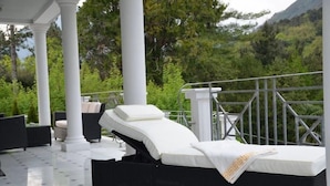 Un espacio lounge al aire libre iluminado por el sol, con una sola silla reclinable y una pequeña mesa, rodeado de columnas blancas y una barandilla de metal con vistas a un paisaje verde y exuberante.