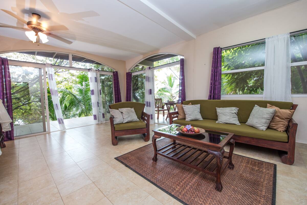 A spacious living room with a ceiling fan, two sofas with cushions, a coffee table, and a glass door leading to a balcony with curtains.