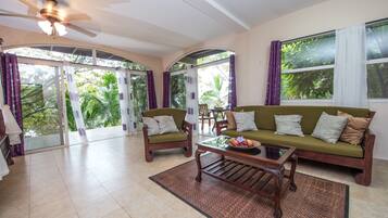A spacious living room with a ceiling fan, two sofas with cushions, a coffee table, and a glass door leading to a balcony with curtains.
