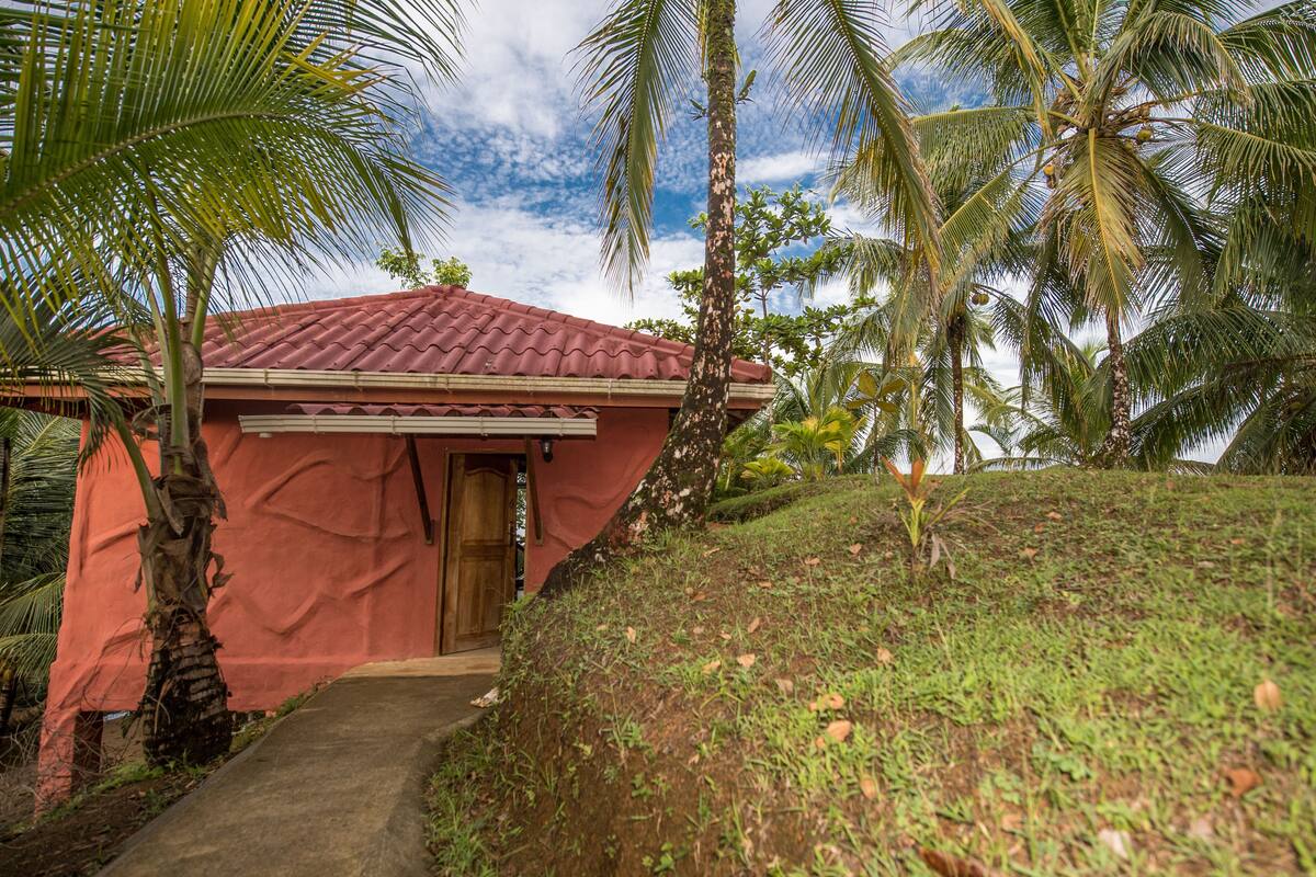 A small, red-roofed building with a wooden door, surrounded by palm trees and a grassy hill.