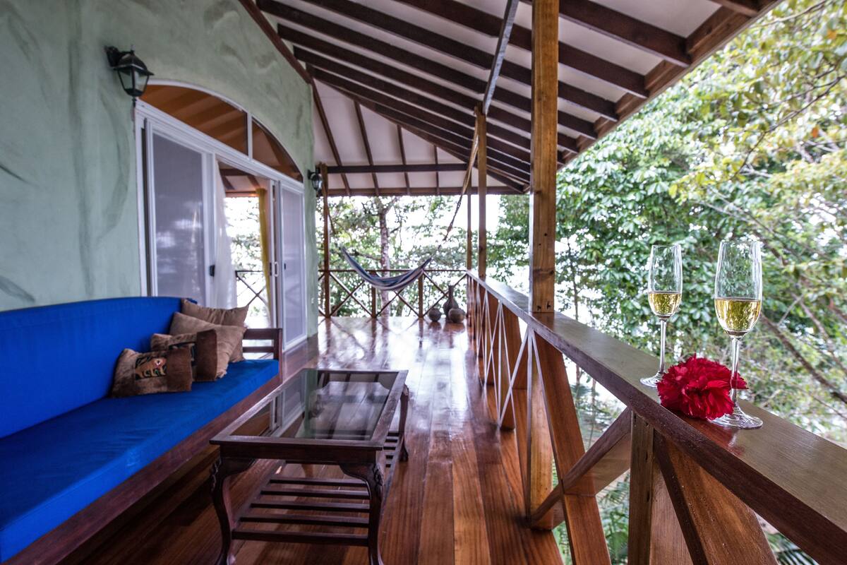 A wooden deck with a blue sofa and a glass table, overlooking a hammock and lush greenery.