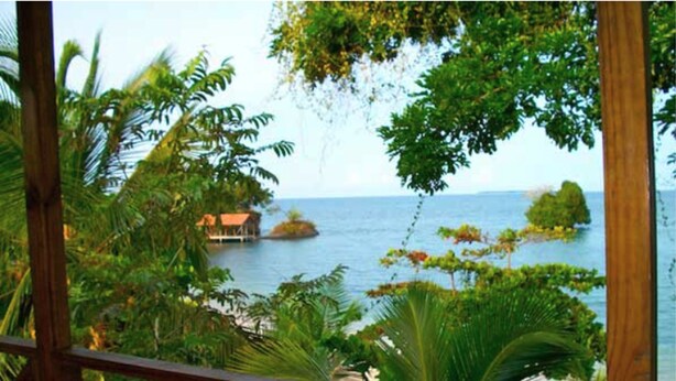 Un pontile di legno con vista su una spiaggia tropicale, una piccola isola e una casa.