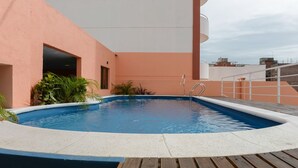 Una piscina en la azotea con un muelle de madera, rodeada por un edificio rosa con un balcón blanco.