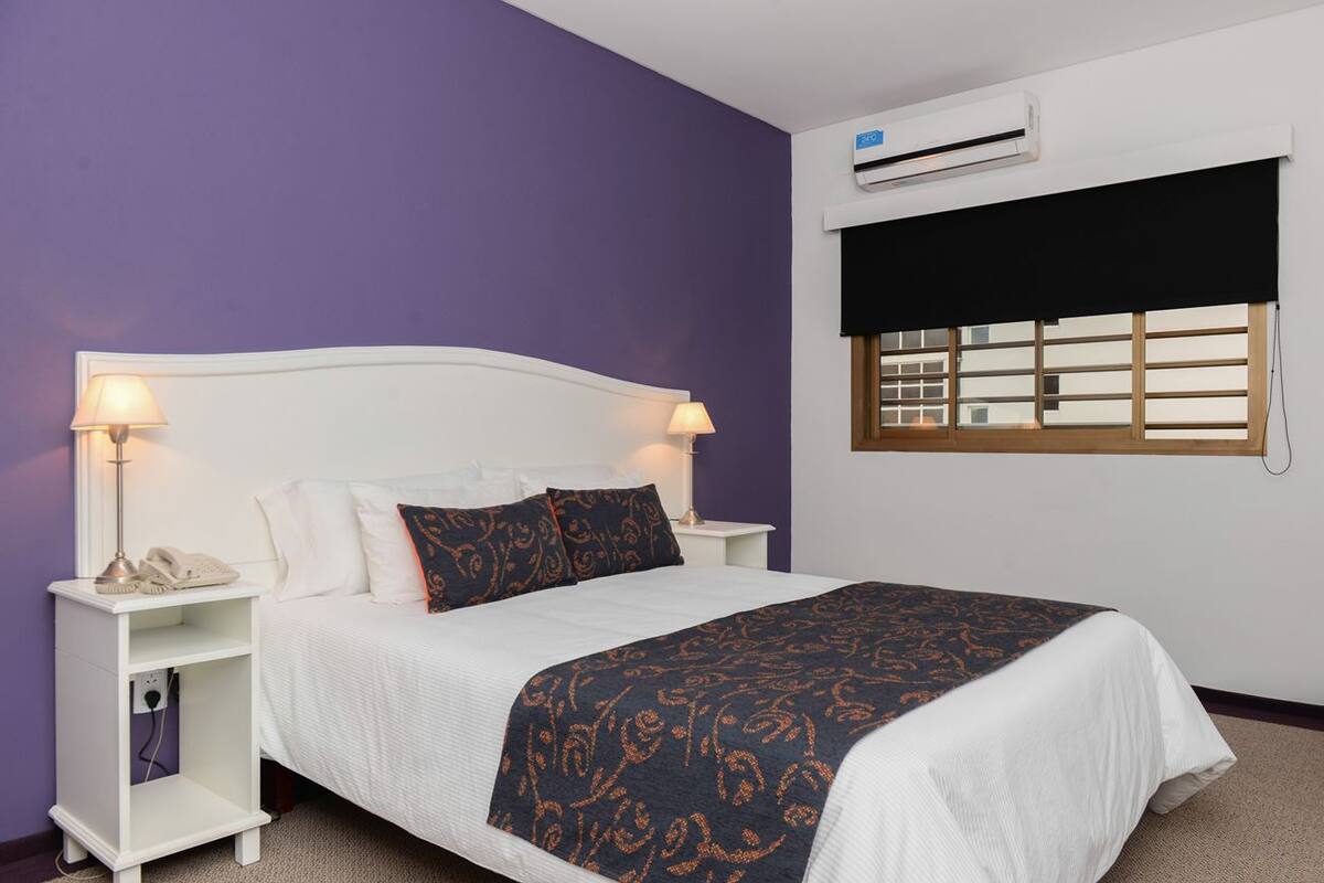 Une chambre à coucher comprenant un lit, deux tables de chevet avec des lampes, une fenêtre avec des stores et un appareil de climatisation.