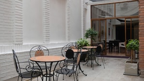 Un patio al aire libre con mesas y sillas redondas, una puerta de vidrio que da a un espacio interior, y plantas en macetas.