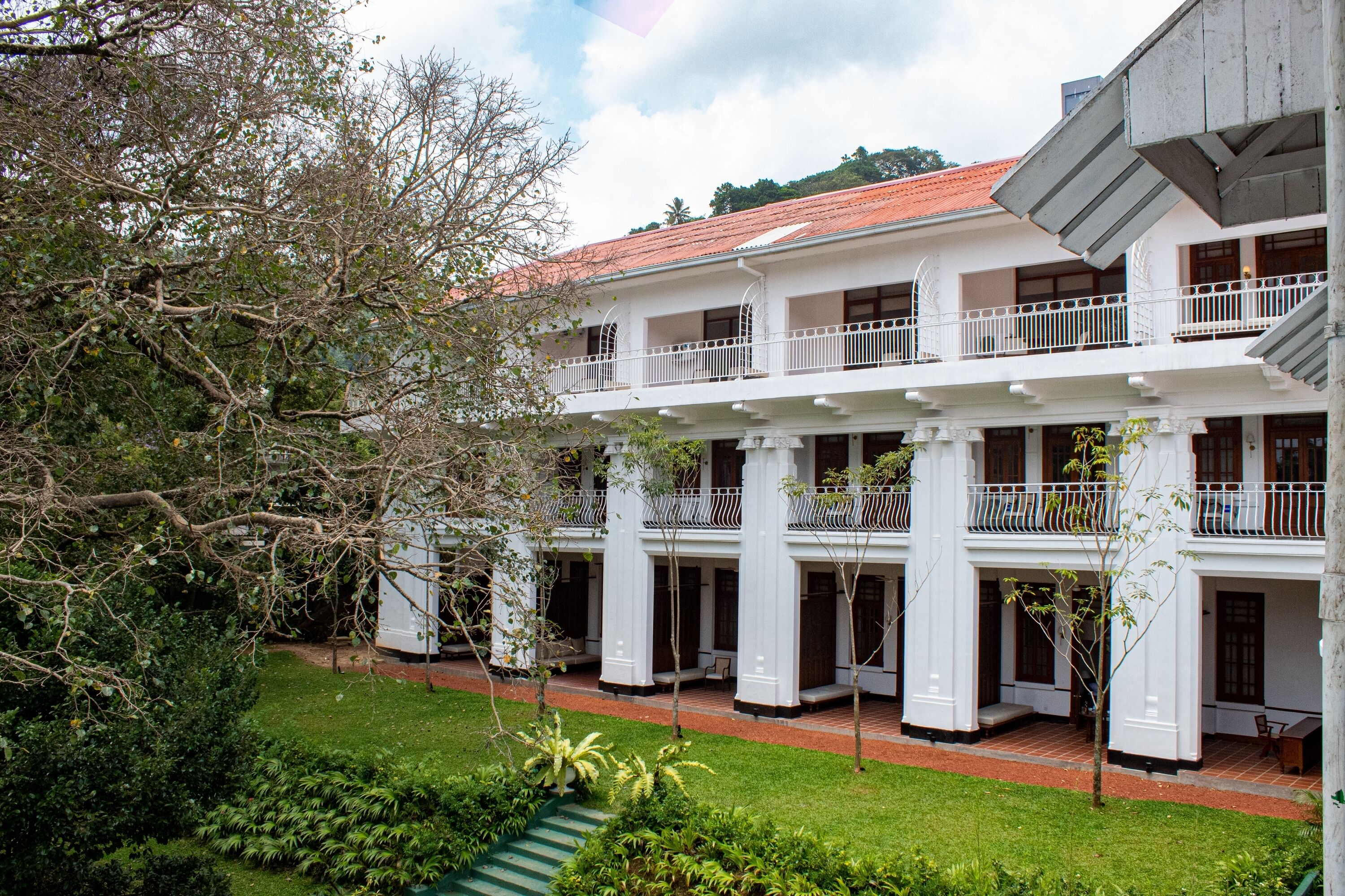 A two-story colonial-style building with a red-tiled roof, white balconies, and a well-maintained garden with green lawns and trees.