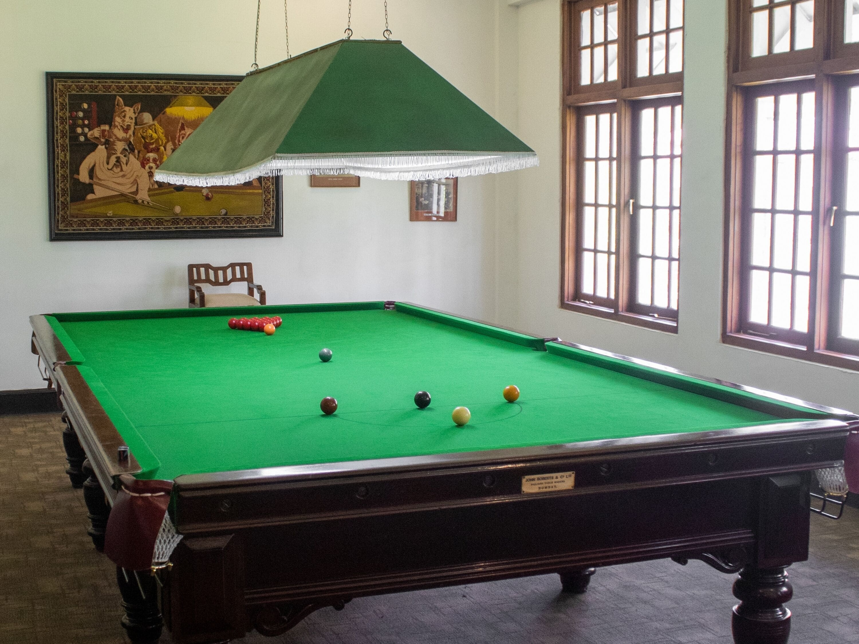 Een biljarttafel met ballen en een keu, een ingelijst beeld aan de muur en een raam met zonwering.