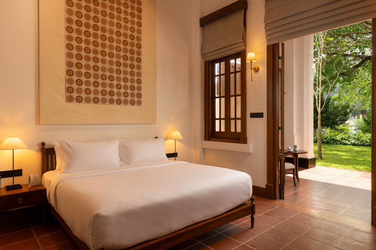 A neatly made bed in a room with a large patterned artwork on the wall, a window with blinds, and a view of a garden outside.