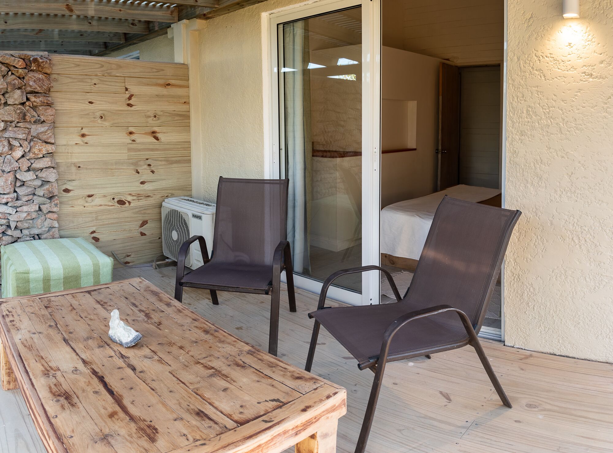 A wooden table with a cushion, two chairs, a stone wall, and a sliding glass door leading to a bedroom.