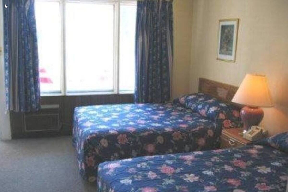 A hotel room with two beds, a window with curtains, a lamp, and a framed picture on the wall.