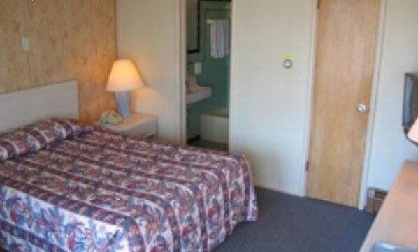 A hotel room with a bed, a nightstand with a lamp, a bathroom visible through an open door, and a television on a stand. - Shiva's Travelers Lodge (Niagara Falls)