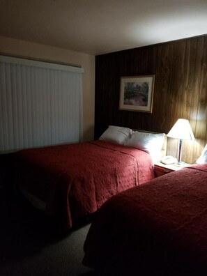 A hotel room with two beds, a nightstand, a lamp, a painting on the wall, and a window with blinds. - Shiva's Travelers Lodge (Niagara Falls)