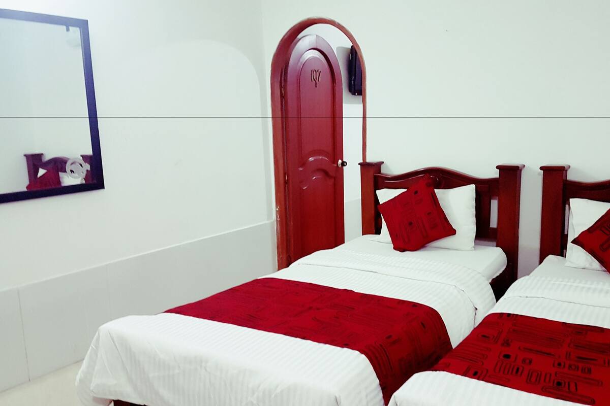 Quarto de hotel com duas camas, cada uma com roupa de cama branca e almofadas vermelhas. Há uma cabeceira de madeira, uma porta vermelha e um espelho na parede.