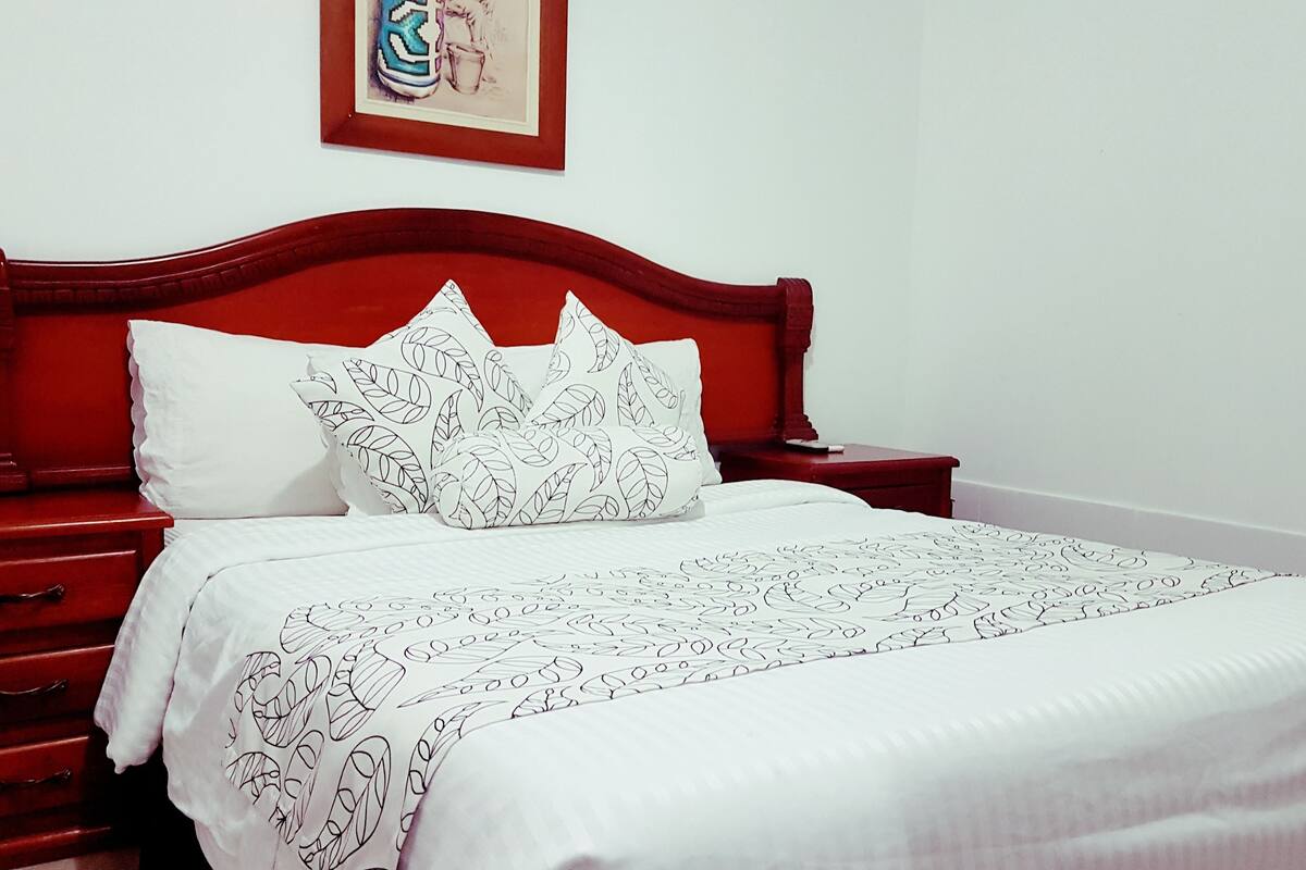 Uma cama bem arrumada com colcha branca e almofadas estampadas com folhas, cabeceira de madeira e mesa de cabeceira vermelha com gavetas.