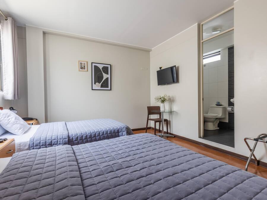 A hotel room with two beds, a desk, a chair, a TV, and a bathroom visible through a glass door.
