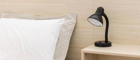 A neatly made bed with a white pillow and a quilted headboard, a black desk lamp on a wooden nightstand, and a plain wall in the background.