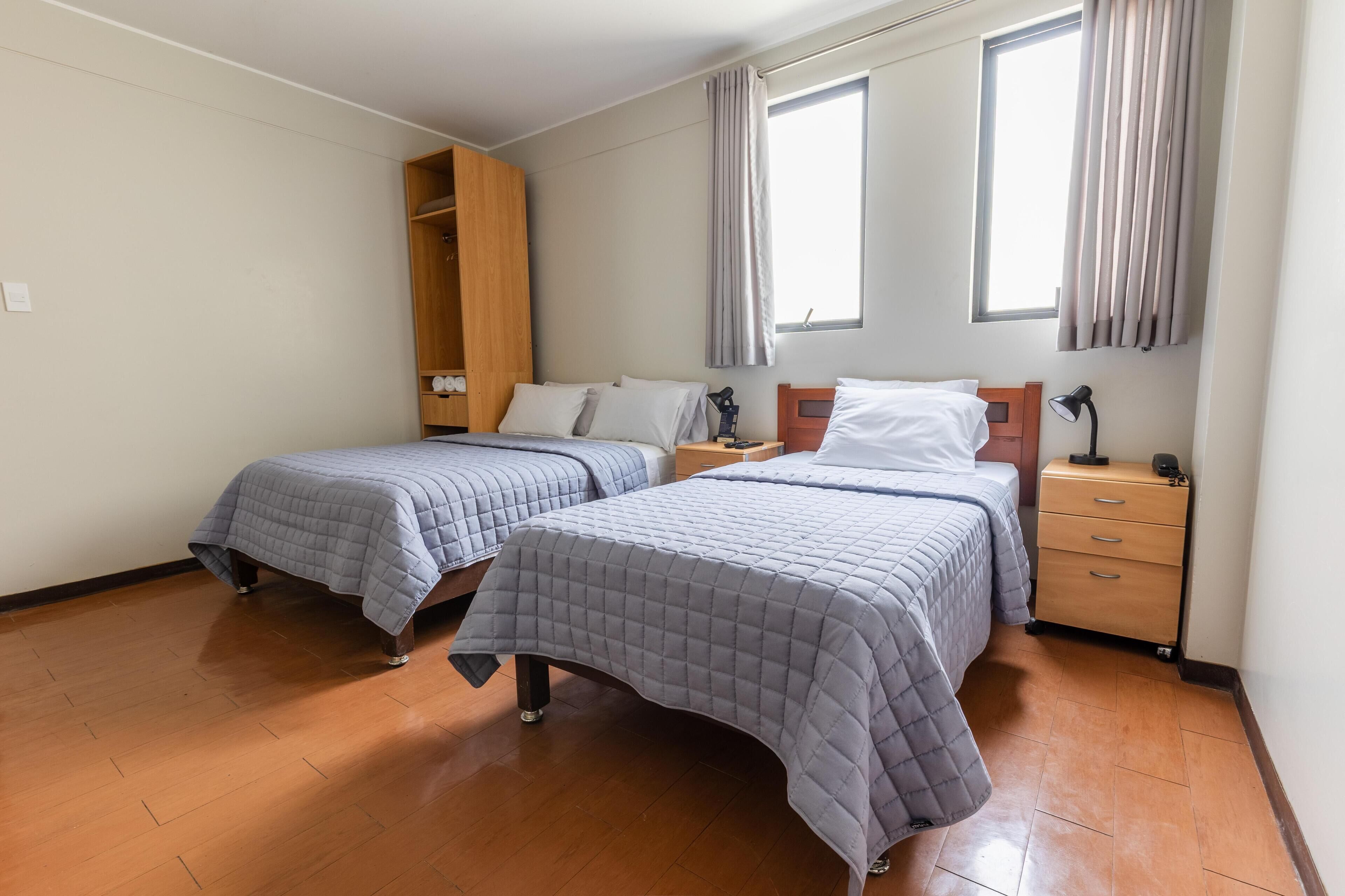 A hotel room with two beds, wooden wardrobe, bedside tables, and a window with curtains.