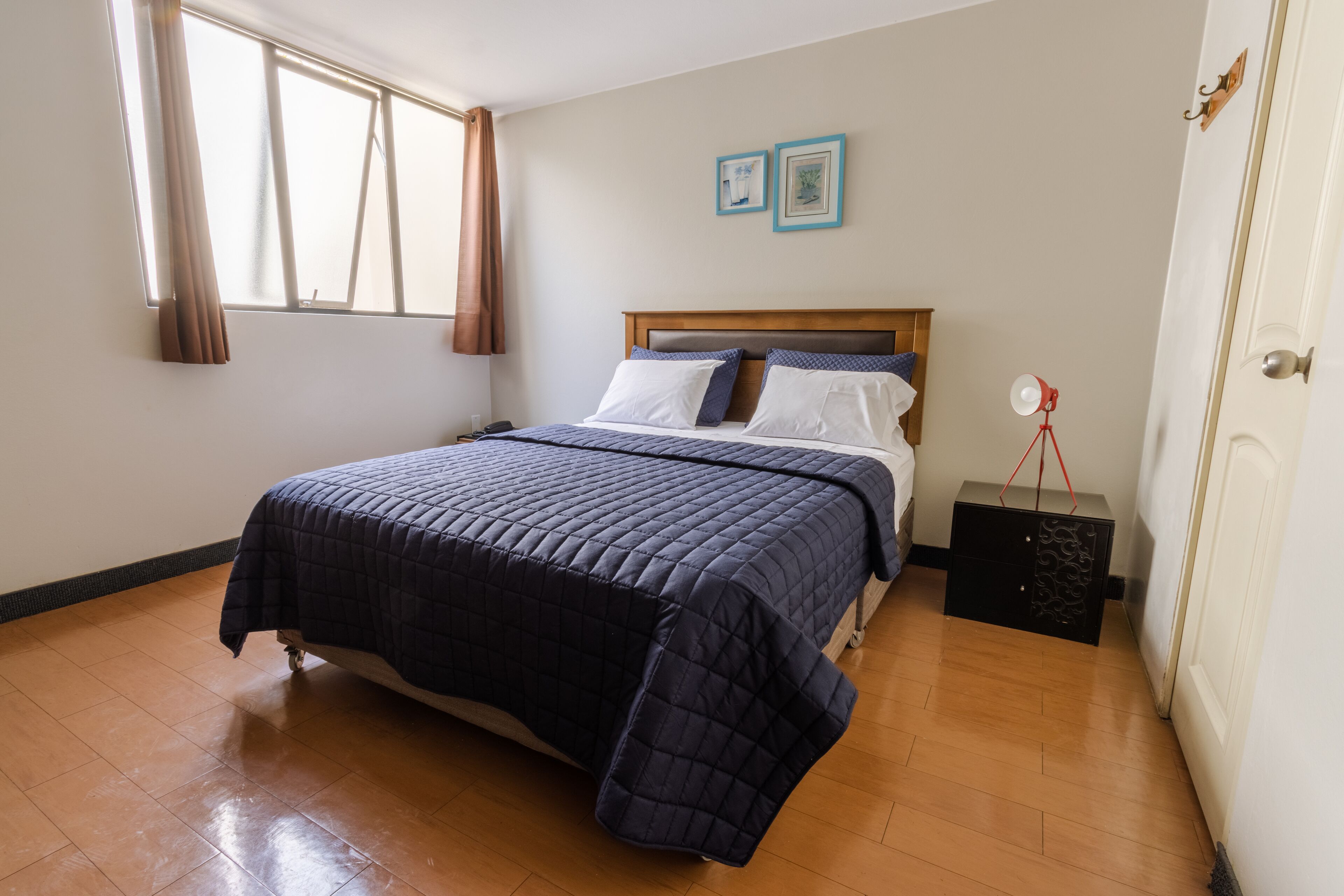 Een slaapkamer met een bed, houten vloeren, een raam met gordijnen en een bedlampje.
