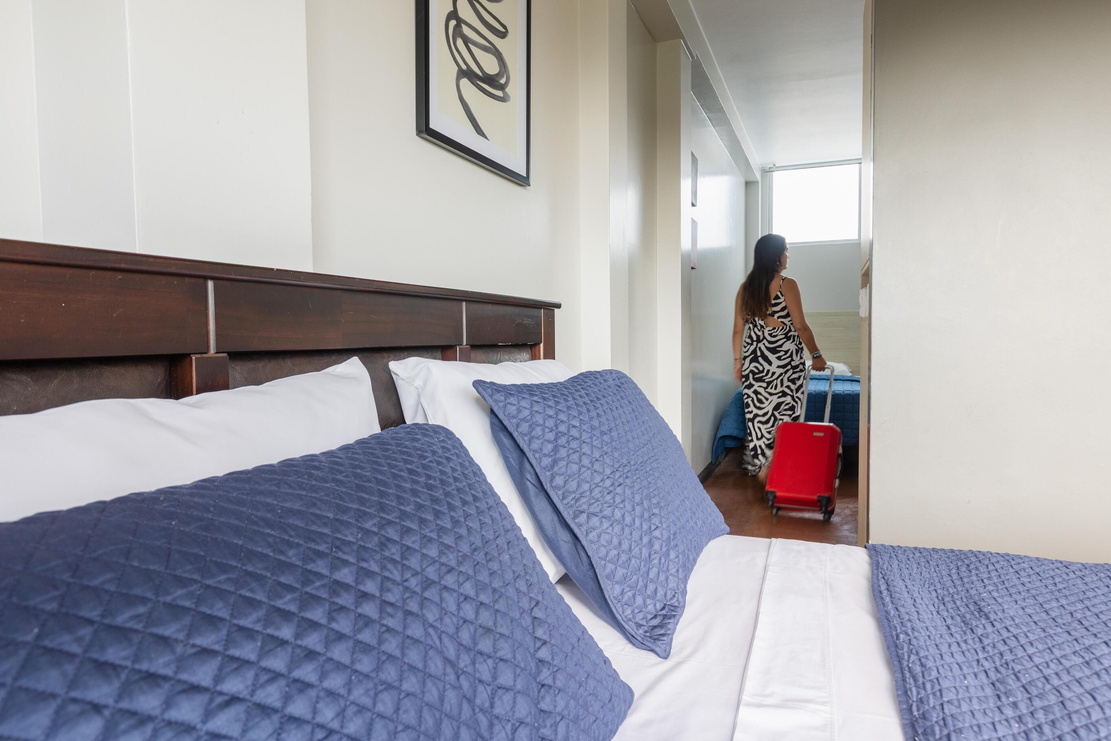 A hotel room with a large bed, a headboard, a framed artwork on the wall, and a view of a hallway with a person and luggage.