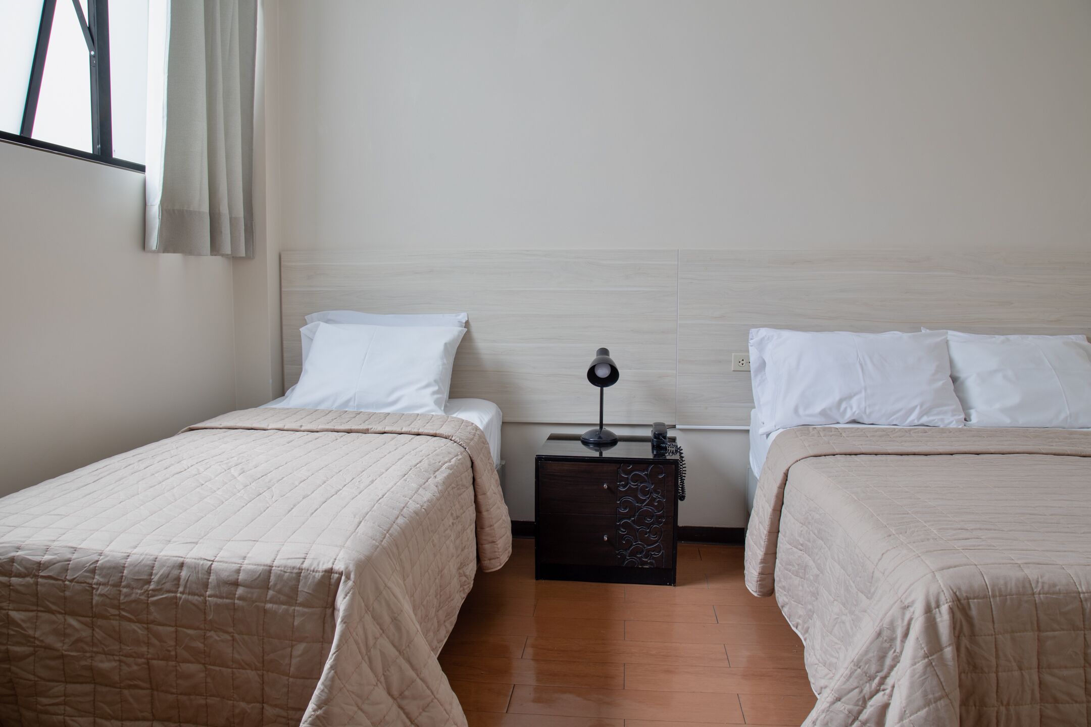 Hotellihuone, jossa on kaksi sänkyä, yöpöytä lampun kanssa ja puulattia.