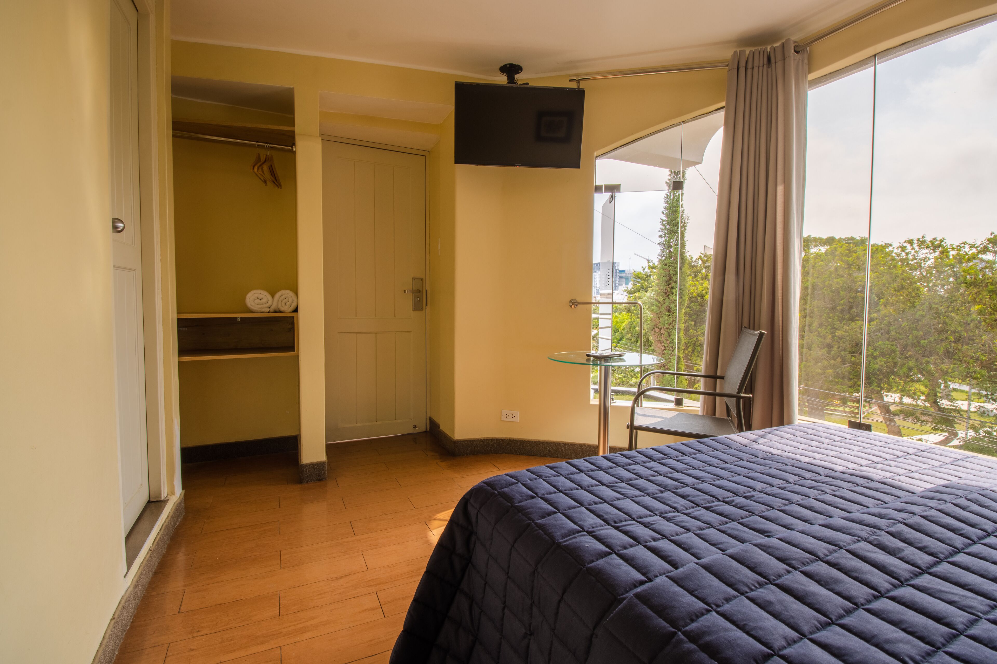 Habitación de hotel con cama, una mesita, una silla, televisión y balcón con vistas a árboles y edificios.