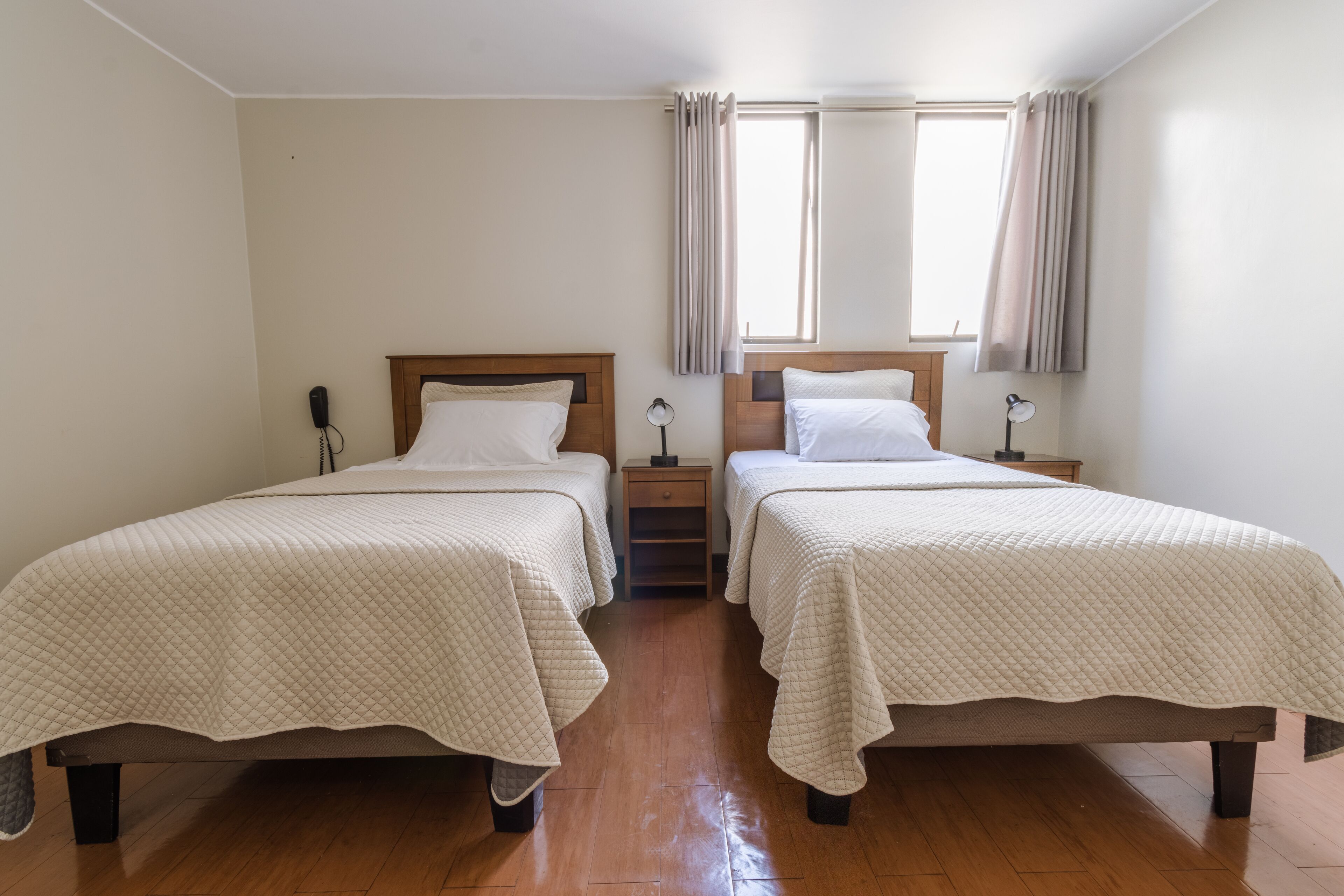 A room with two beds, wooden headboards, a small nightstand with a lamp, and a window with curtains.