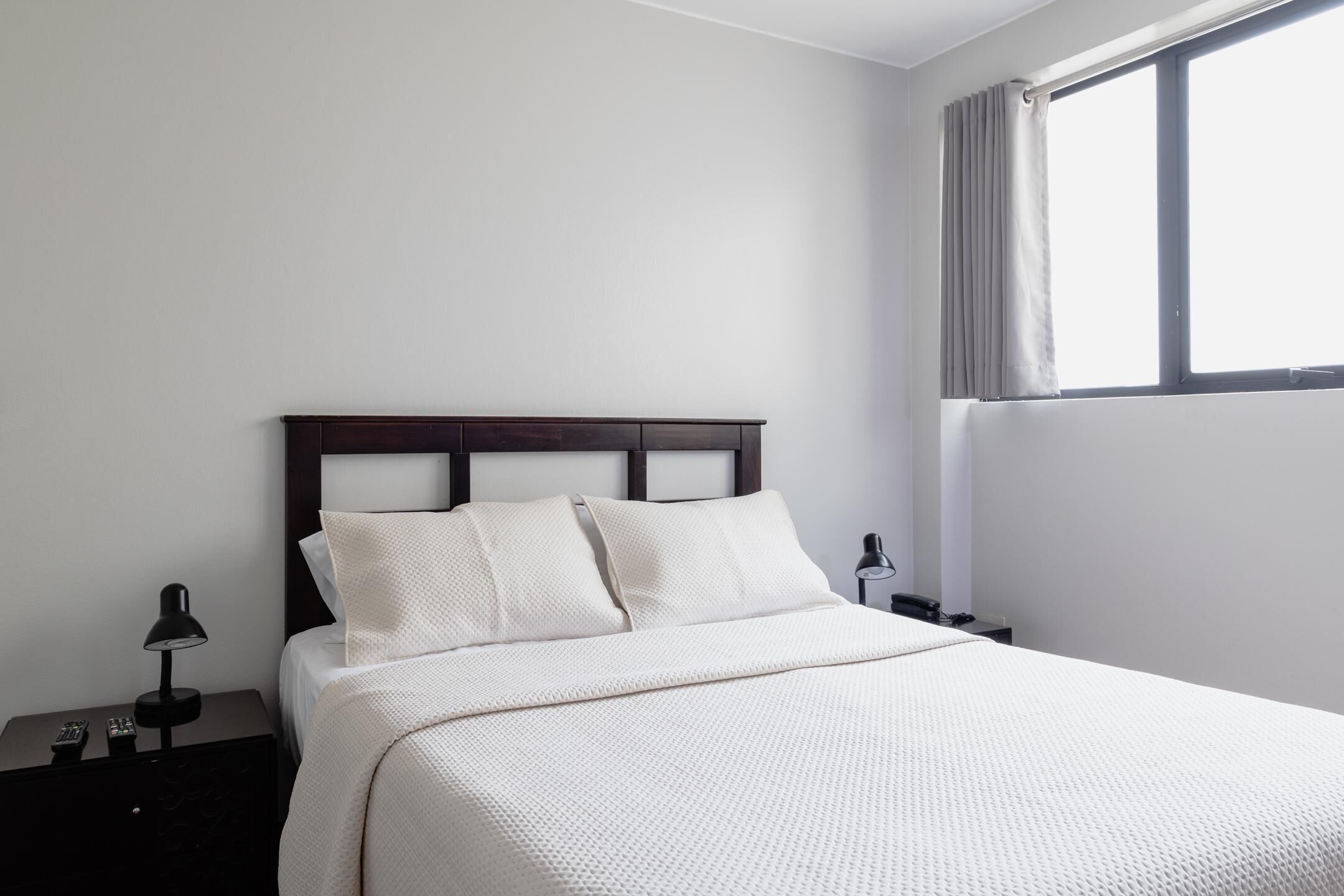 A neatly made bed with a white comforter and pillows, a dark wooden headboard, a nightstand with a lamp, and a window with curtains.