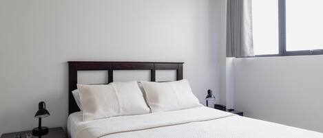 A neatly made bed with a white comforter and pillows, a dark wooden headboard, a nightstand with a lamp, and a window with curtains.