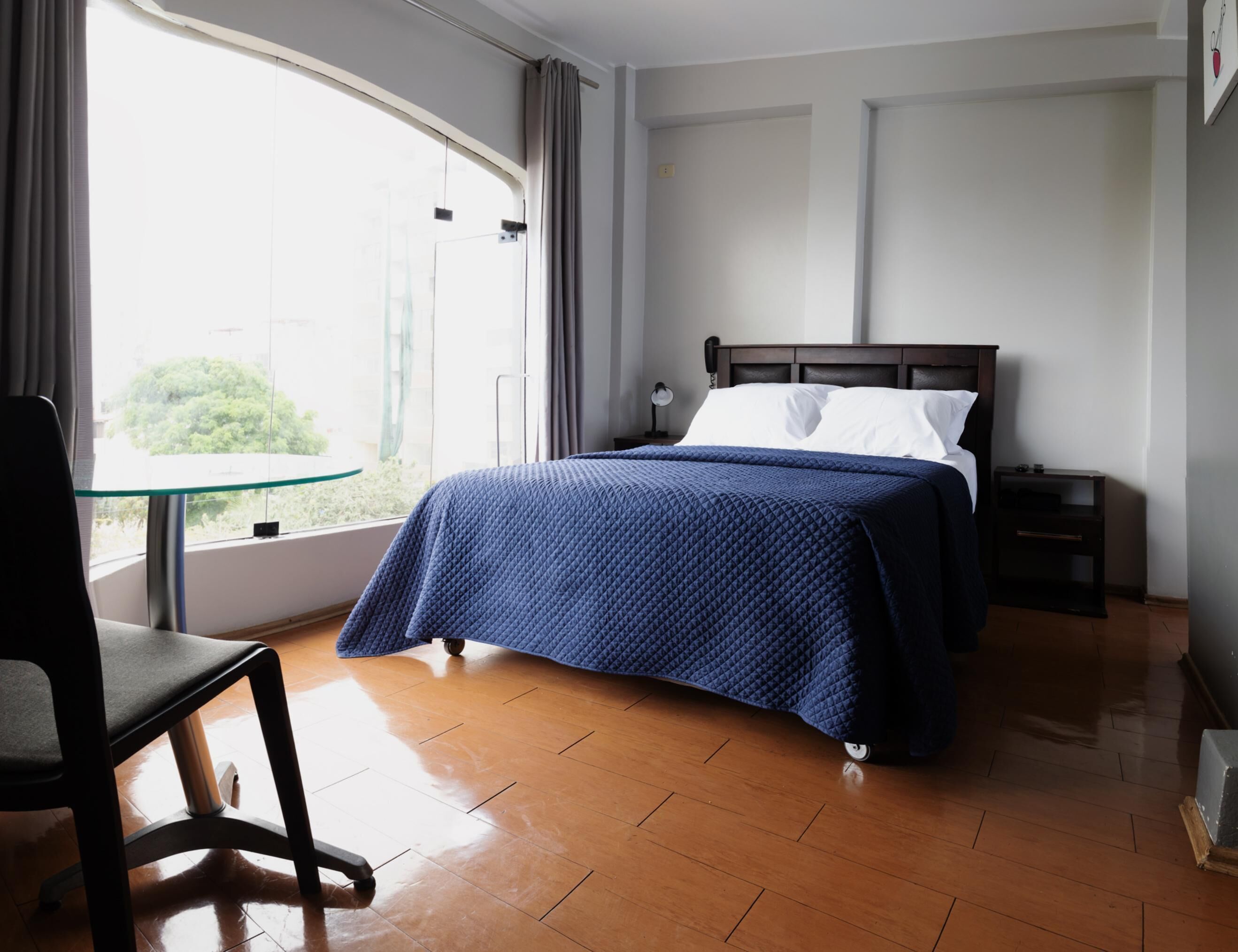 A hotel room with a bed, a chair, a nightstand, a bedside table, a window with curtains, and a view of greenery outside.