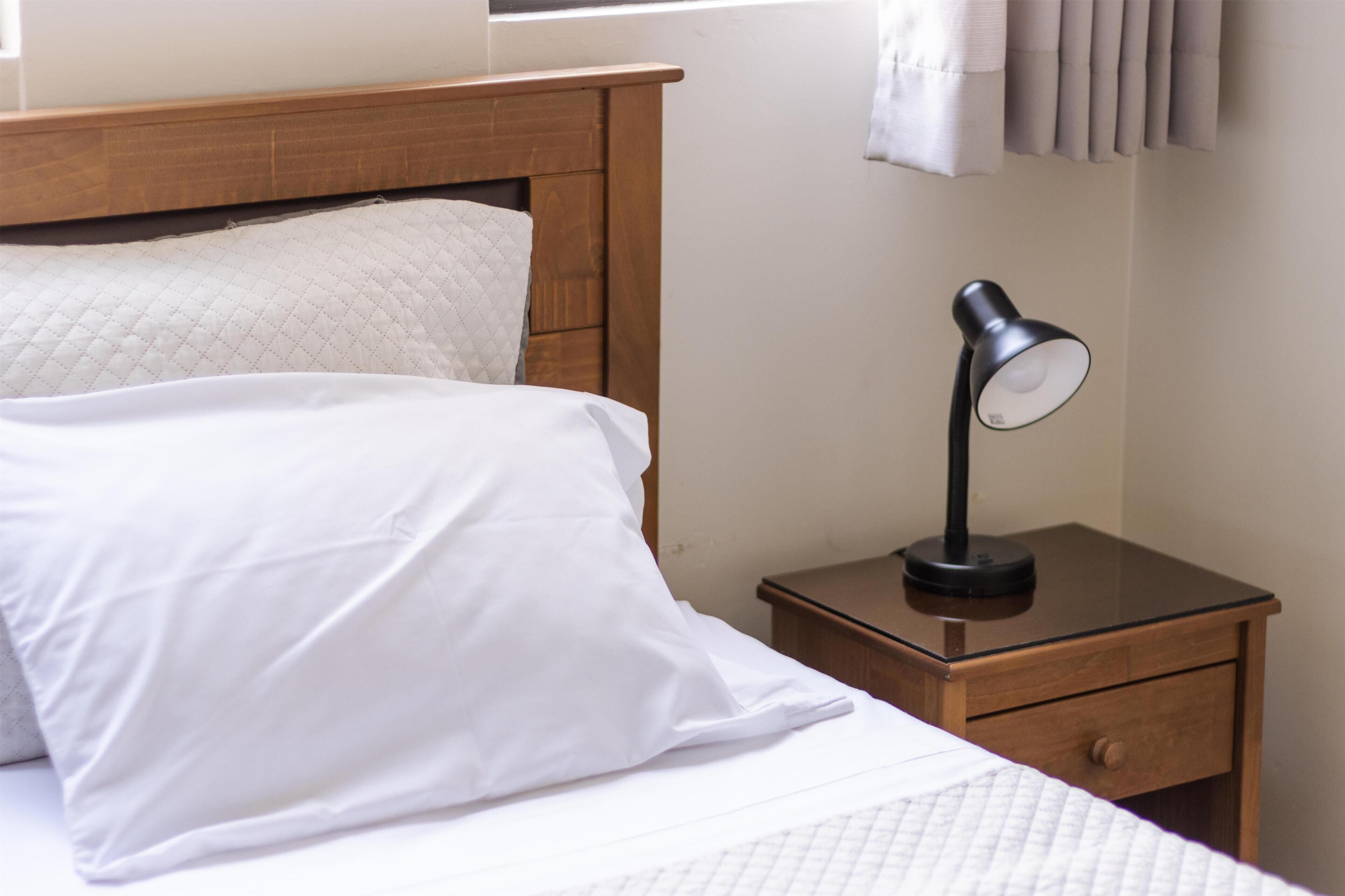 A neatly made bed with white bedding and a wooden headboard. A small wooden nightstand with a black lamp is beside the bed.