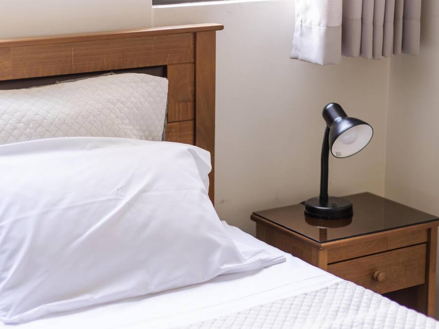 A neatly made bed with white bedding and a wooden headboard. A small wooden nightstand with a black lamp is beside the bed.