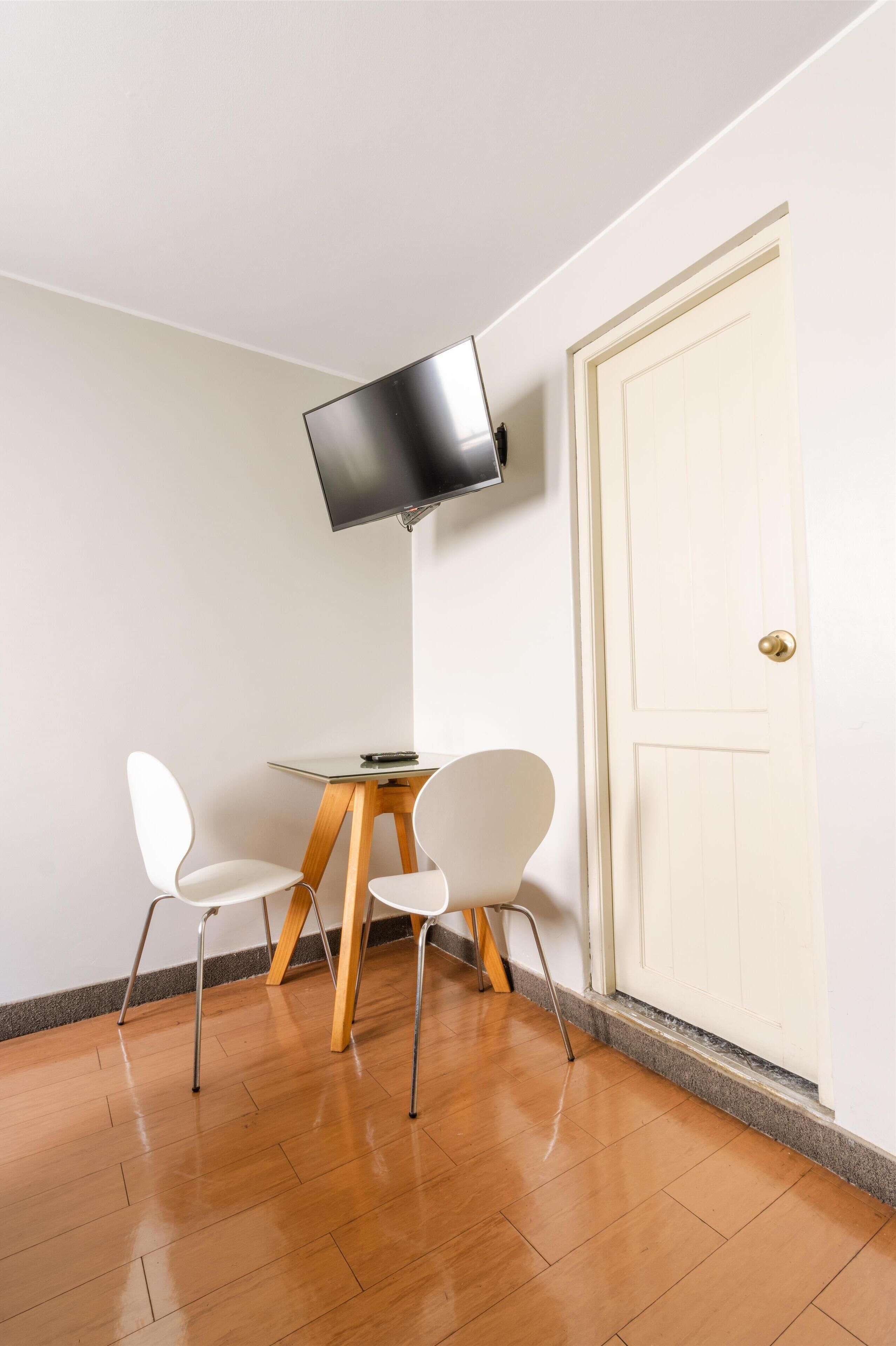 Ein Eckzimmer mit Holzboden, einer weißen Tür und einem kleinen Tisch mit zwei weißen Stühlen. Ein Flachbildfernseher ist an der Wand oberhalb des Tisches angebracht.