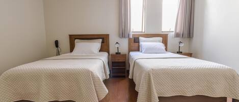 A room with two beds, wooden headboards, a nightstand, a lamp, and a window with curtains.