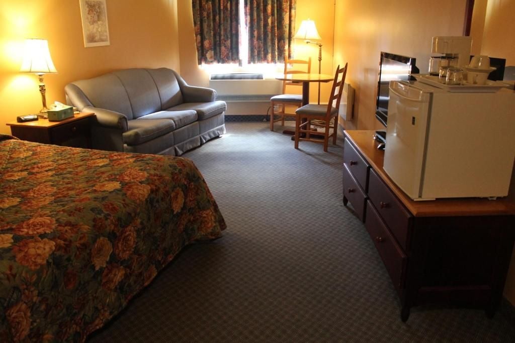 Habitación de hotel con una cama, un sofá, un escritorio con refrigerador, una lámpara y una ventana con cortinas.