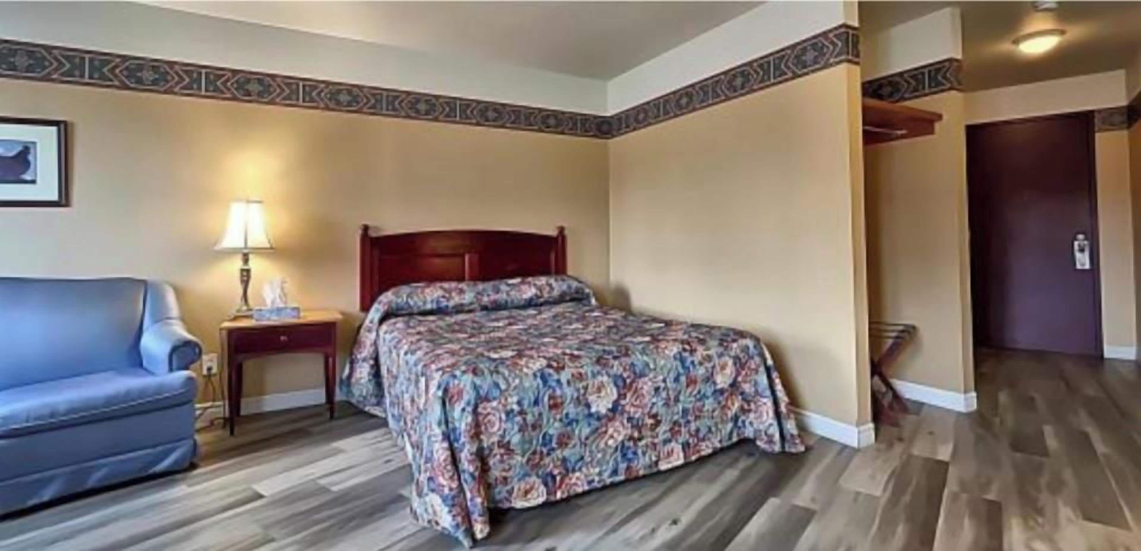 Habitación de hotel con cama, mesita de noche, lámpara, sofá azul y suelo de madera.